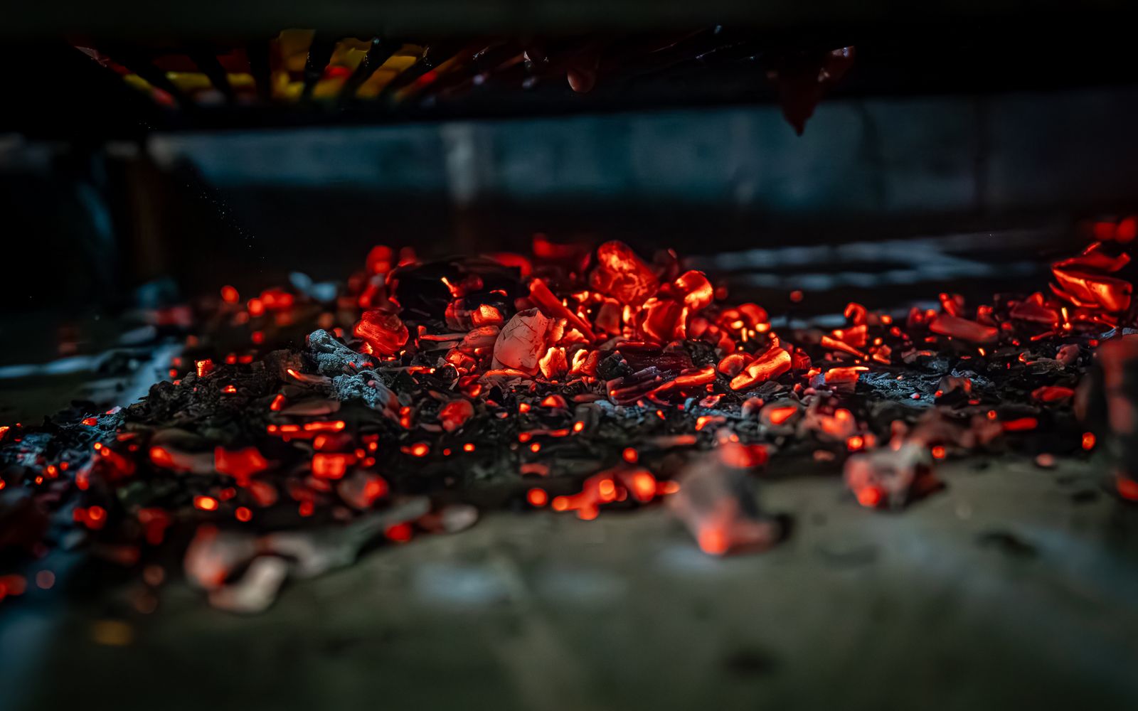 Photo d’illustration — braises et flammes basses sous une grille, très sombre, orangé profond